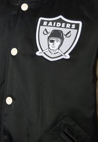 Μαύρο μπουφάν με κέντημα λογότυπου Raiders. Κουμπιά τύπου σνάπ σε ελεφαντόδοντο χρώμα, με υφασμάτινη υφή και κεκλιμένη λεπτομέρεια τσέπης.