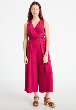Kobieta ubrana w bezrękawnikowy, głęboko różowy kombinezon o szerokich nogawkach z dekoltem w serek i wzorzystym paskiem w talii, w połączeniu z brązowymi sandałami na obcasie, stojąca na białym tle.
