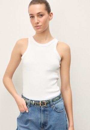 Mlada ženska z zglajenimi lasmi, oblečena v belo majico brez rokavov in modre jeans hlače z vzorčastim pasom, stoji pred enobarvnim ozadjem.