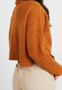 Pomarańczowy sweter dziany z prążkowaną teksturą i wysokim kołnierzem. Dobranie do kremowych spodni z bocznymi kieszeniami.
