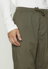Gros plan d'une personne portant un pantalon à cordon vert olive et une chemise blanche à manches longues, la main reposant à côté.