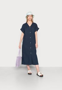 Vestido azul-marinho, até aos joelhos, com botões, decote em V, mangas curtas e um bolso no peito. Combinado com sandálias brancas e uma bolsa roxa.