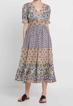 Kvinde iført en multifarvet midi kjole med blomster- og geometrisk mønster, korte ærmer og sorte flade sandaler med åben tå.