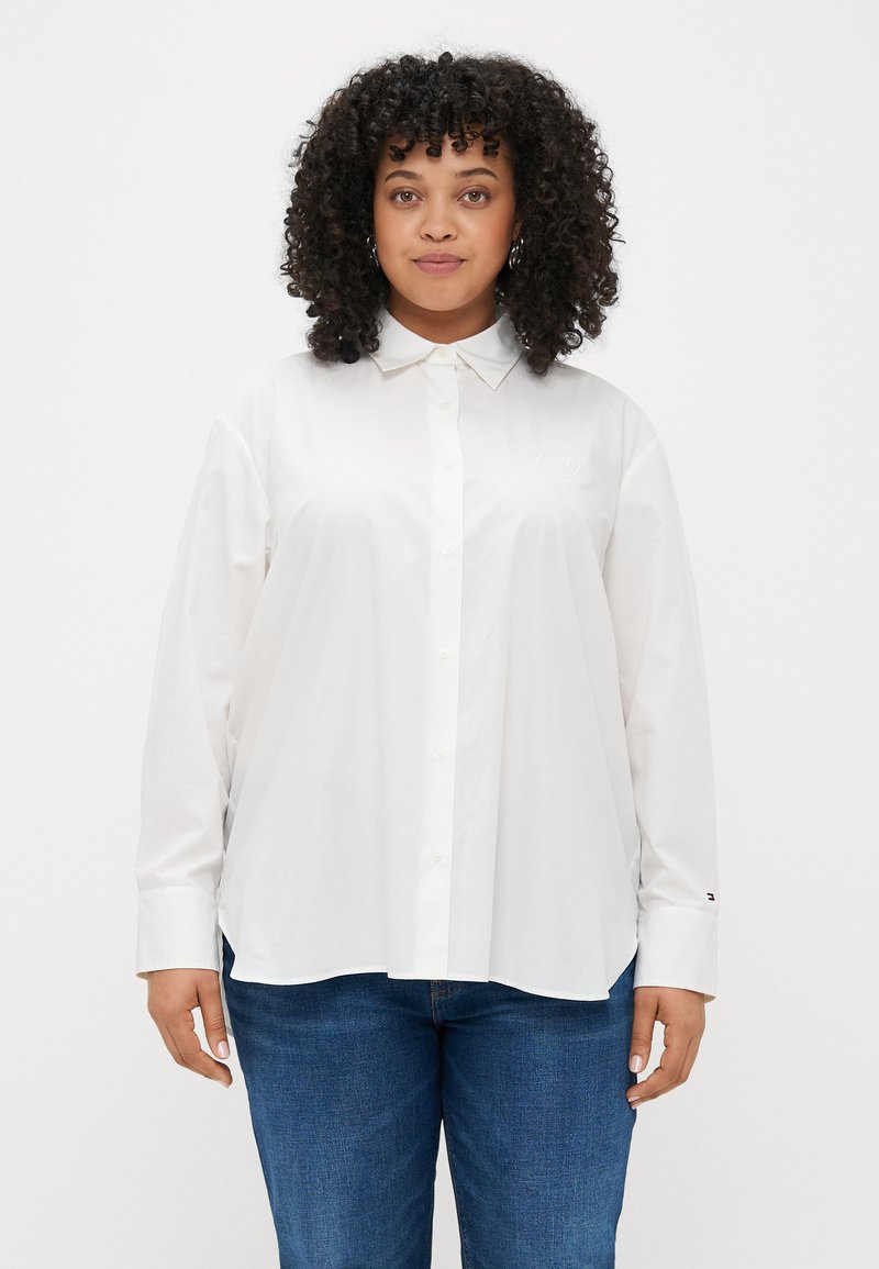 White long-sleeve button-up shirt made of smooth fabric, featuring a classic collar and a subtle logo detail on the chest. Paired with blue jeans.