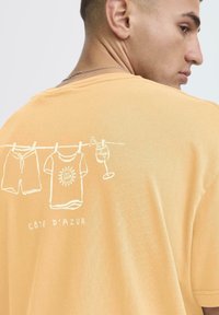 Krátký rukáv, oranžové bavlněné tričko s bílým liniovým uměním oblečení a nápoje. Text zní "CÔTE D’AZUR" pod designem.