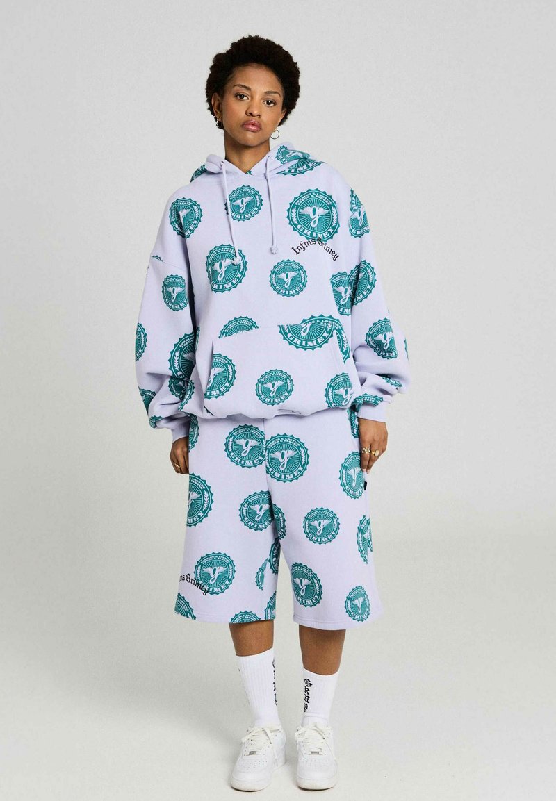 Mujer con sudadera lavanda extragrande y pantalones cortos a juego de largo hasta la rodilla, con patrones de logotipos circulares verdes, calcetas blancas y zapatillas deportivas.