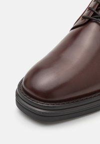 Chaussure en cuir marron avec un bout arrondi, présentant une texture lisse et une semelle en caoutchouc noire contrastante. Couture détaillée le long du bord.