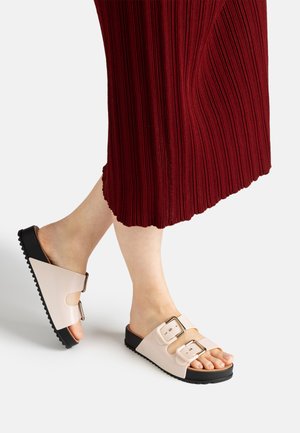 Feet wearing beige slide sandals with two buckles and black soles, paired with a long red ribbed skirt.