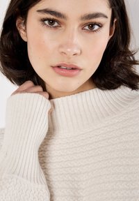 Kremowy sweter z wysokim ściągaczem i teksturowanym wzorem dzianiny. Posiada długie rękawy z ściągaczami i miękkie, dziane wykończenie.