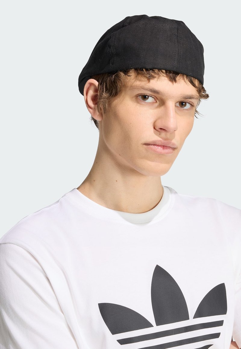 Junger Mann mit schwarzer Kappe und weißem T-Shirt mit großem schwarzem Adidas-Logo, der leicht zur Seite blickt vor schlichtem Hintergrund.