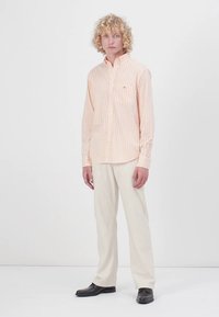 Camicia a righe arancione chiaro e bianca, in tessuto di cotone, maniche lunghe, taschino sul lato sinistro del petto. Abbinata a pantaloni crema e scarpe nere.
