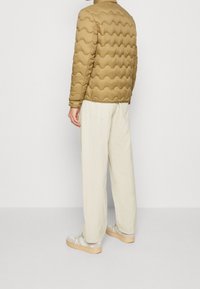 Hombre vestido con una chaqueta acolchada de color marrón claro, pantalones sueltos color blanco roto y zapatillas beige, de pie contra un fondo blanco liso.