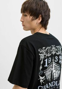 Sort bomulds t-shirt med en dekorativ hvid grafik på ryggen, der viser en bygning og ornamentalt design, kortærmet.