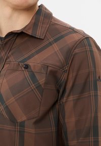 Bruin geruite shirt met zwarte en oranje accenten. Bevat een knoopsluiting op de borstzak, een puntige kraag en een gestructureerde stof.