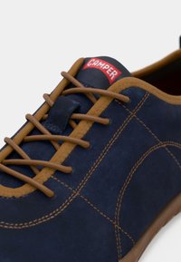 Chaussure en daim bleu marine avec des lacets et des coutures marron, arborant une étiquette de logo Camper en haut. Bout rond et semelle plate en caoutchouc.