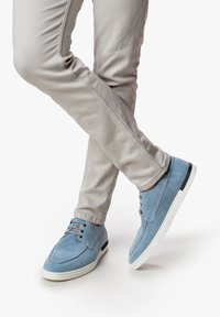 Zapatos de gamuza azul claro con cierre de cordones, suela blanca y sutiles acentos oscuros, combinados con pantalones ajustados gris claro.