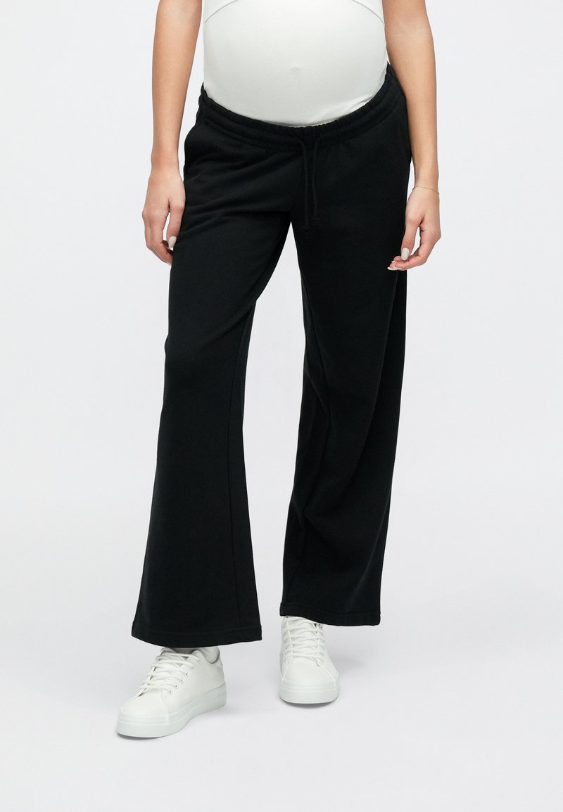 Gravid person iført sorte, vidde bukser til gravide og hvide sneakers, stående mod en ensfarvet hvid baggrund.