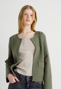 Ελαιόλαδο πράσινο μπουφάν με στρογγυλή λαιμόκοψη, μία τσέπη και μαλακή υφή, σε συνδυασμό με ένα ανοιχτό μπεζ ριμπ κορμάκι και σκούρα τζιν.