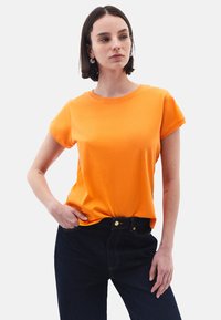 Tričko s krátkým rukávem a kulatým výstřihem v živé oranžové barvě z bavlny. Má hladkou strukturu a volný střih, kombinované s tmavými denimoovými džíny.