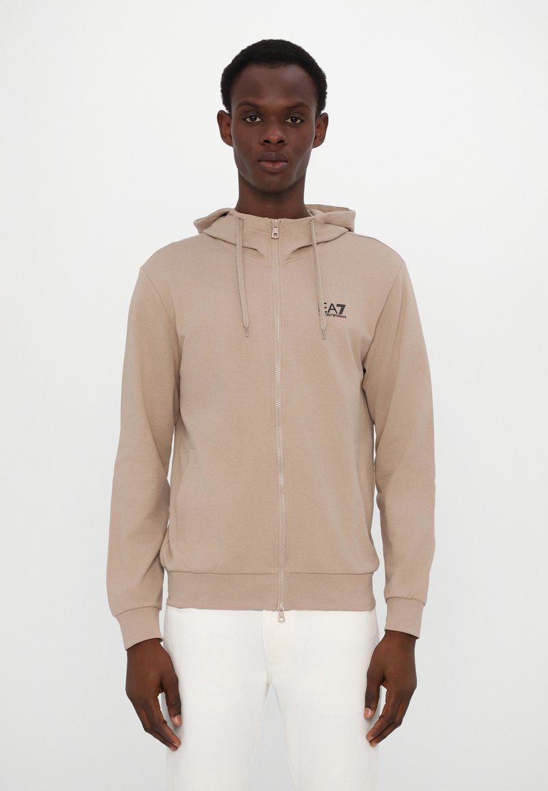 Sudadera con cremallera en color beige, con capucha ajustable mediante cordones, cierre de cremallera en el frente y logo negro en el pecho. Tejido suave con una textura lisa.