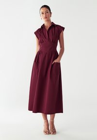 Abito bordeaux con scollatura a colletto, maniche a cappuccio e due tasche sovradimensionate. Tessuto liscio, lunghezza al ginocchio e aderente in vita.