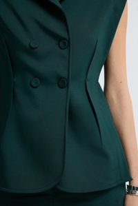 Bezrukávový tmavozelený dvouřadý blejzr se čtyřmi zakrytými knoflíky, který je na sobě, přičemž je vidět část paže a zápěstí s stříbrným náramkem.