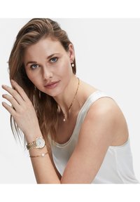 Gouden polshorloge met een witte wijzerplaat, gekoppelde armband. Model draagt een gouden ketting met een parel hanger en bijpassende oorbellen.