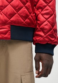 Κόκκινο καπιτονέ μπουφάν bomber με ναυτικές ριγέ μανσέτες. Το ύφασμα έχει γυαλιστερή επιφάνεια και οι μανσέτες διαθέτουν υφασμάτινο, πλεγμένο σχέδιο.