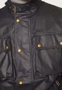 Svart vattentät jacka med hög krage, två framfickor med mässingsknappar och subtil quiltning på axlarna.