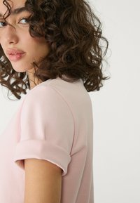 Lyse pink, kortærmet skjorte med rund krave og oprullede manchetter, lavet af blødt stof. Designet har en afslappet pasform.