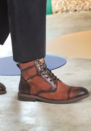 Botas de cuero marrón con cordones hasta el tobillo, combinadas con pantalones negros sobre un suelo de concreto claro con alfombrillas circulares azules y beige.