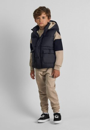 Marineblaue Steppweste mit fellgefütterter Kapuze, zwei Fronttaschen; kombiniert mit beigem Sweatshirt und passenden Jogginghosen; schwarze Sneakers.