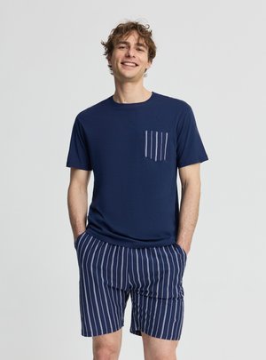 Giovane uomo sorridente con le mani nelle tasche, indossa una maglietta blu navy con tasca a righe e pantaloncini a righe coordinati su sfondo semplice.