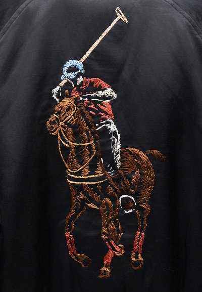 Čierna tkanina s detailnou výšivkou poloprofesionálneho hráča na hnedom koni, ktorý má modrú prilbu, červenú košeľu a červené čižmy.