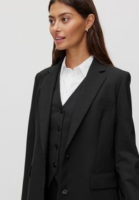 Zwarte blazer met een scherpe revers, getailleerde pasvorm, één borstzak en twee zijzakken. Draag het over een wit overhemd met kraag en een zwart vest.