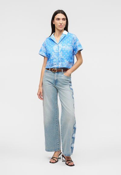 Γυναίκα που φοράει μπλε κοντομάνικο πουκάμισο με σχέδια, ανοιχτόχρωμα τζιν παντελόνια με φαρδιές μπότες και πλευρικά λουλουδάτα σχέδια, καφέ ζώνη και μαύρα πέδιλα με τακούνι, όρθια.