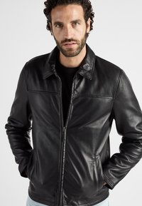 Zwarte leren jas met een ritssluiting aan de voorkant, puntige kraag en twee zijzakken. Gladde textuur, aansluitend ontwerp en zichtbare stikseldetails.