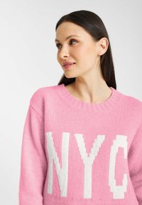 Różowy sweter z dzianiny z białym napisem "NYC". Okrągły dekolt z prążkowanym wykończeniem, miękka tekstura i luźny krój.