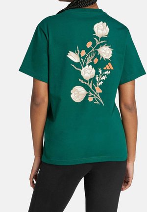 Zielona koszula z krótkim rękawem z dużym białym i pomarańczowym kwiatowym wzorem na plecach, noszona przez osobę z zaplecionymi włosami.