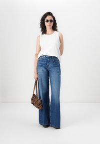 Top branco sem mangas, jeans flared azuis de cintura alta, óculos de sol pretos, e uma mala de ombro castanha, de pé contra um fundo branco.