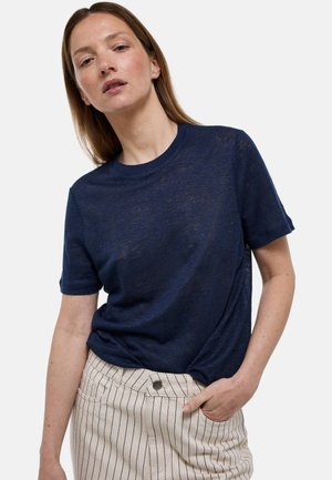 Femme aux longs cheveux châtain clair portant une chemise bleu marine à manches courtes et un pantalon beige à fines rayures, main dans la poche, fond neutre.