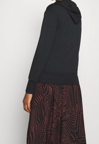 Sweat à capuche noir à manches longues et poignets côtelés, associé à une jupe fluide présentant un motif abstrait rouge et noir.