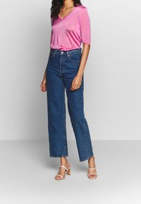 Růžové tričko s výstřihem do V a krátkými rukávy, kombinované s modrými džíny s vysokým pasem a rovnými nohavicemi. Modelka má na sobě světle růžové sandály na podpatku.