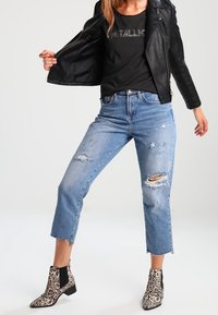 Musta nahkainen moottoripyörätsai, musta graafinen t-paita, kuluneet vaaleansiniset farkut, joissa on repaleinen helma, ja eläinkuosilliset nilkkurit.