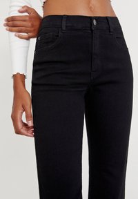 Pantalones de mezclilla negra con un diseño ajustado. Las características incluyen bolsillos laterales, cierre de botón y una textura suave.