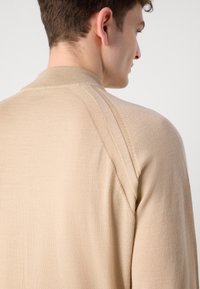 Beige strikkegenser med ribbestrikket krage, innsydd skuldersøm og subtile horisontale sømdetaljer på ermene.
