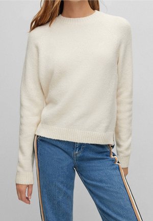 Femme portant un pull en maille crème et un jean bleu avec des bandes latérales beige et noire sur un fond gris uni.