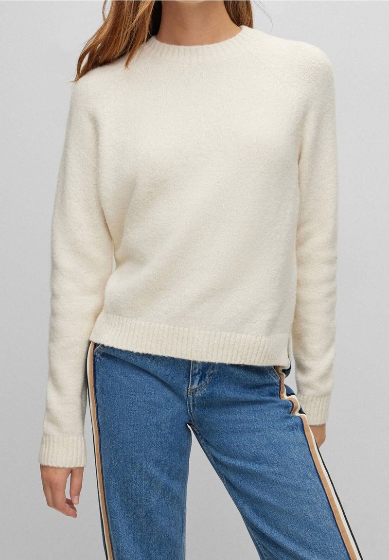 Femme portant un pull en maille crème et un jean bleu avec des bandes latérales beige et noire sur un fond gris uni.