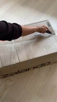 Kartonowe pudełko z oznaczeniem Zalando, zaplombowane przezroczystą taśmą, otwierane nożem tapicerskim na jasnym drewnianym blacie.