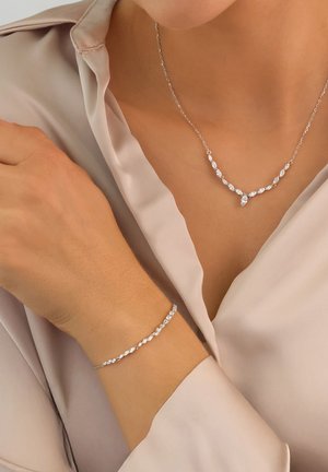 Silver necklace and bracelet feature sparkling clear gemstones in marquise and round shapes, set on delicate chains. Worn with a silky top.